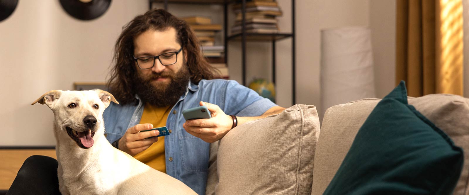 Man paying for something with a card on his smart phone while his dog sits next to him on the couch