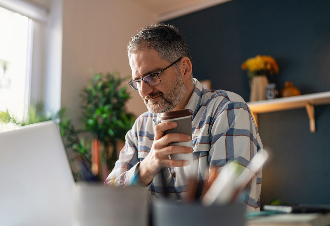 Man smiling while drinking coffee and work on his laptop inside